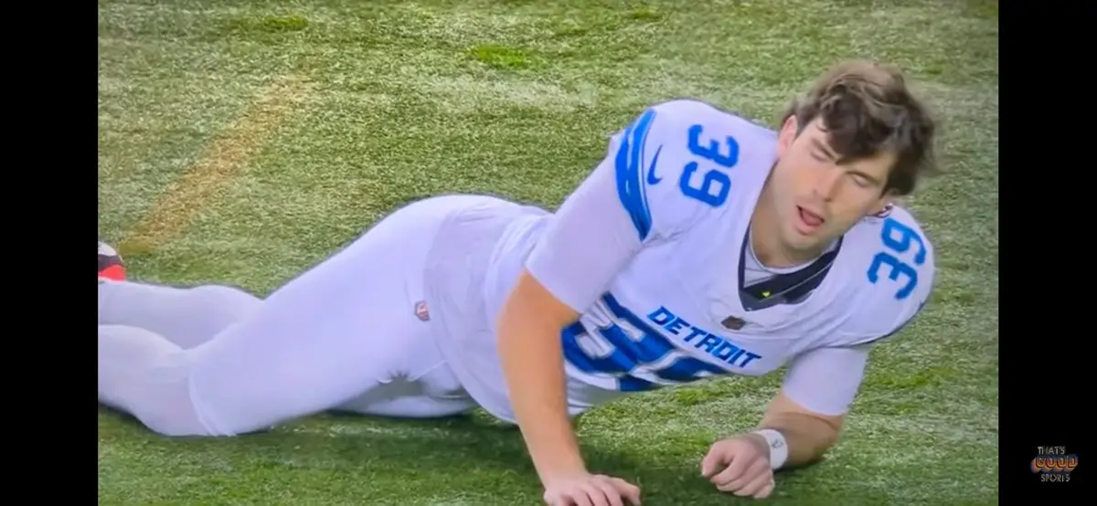 a football player in a white Detroit Lions jersey with number 39 is lying on his stomach on a green field, looking up with a…