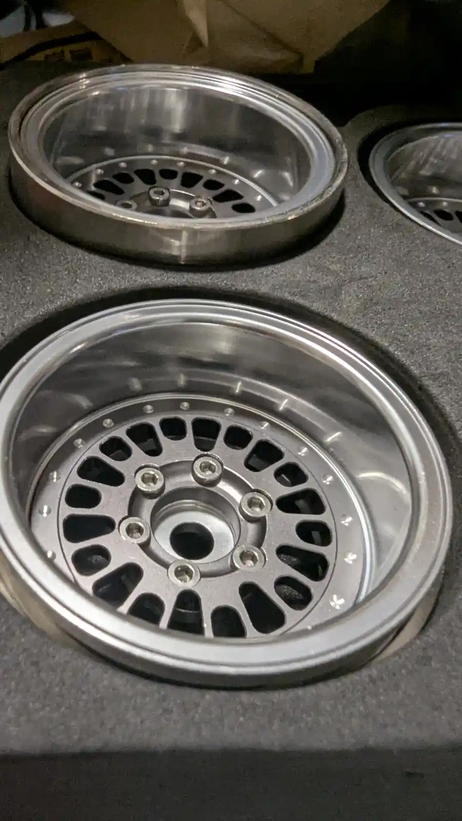 The image shows two shiny, silver, aftermarket car wheels with a multi-spoke design, sitting in a protective foam-lined box…