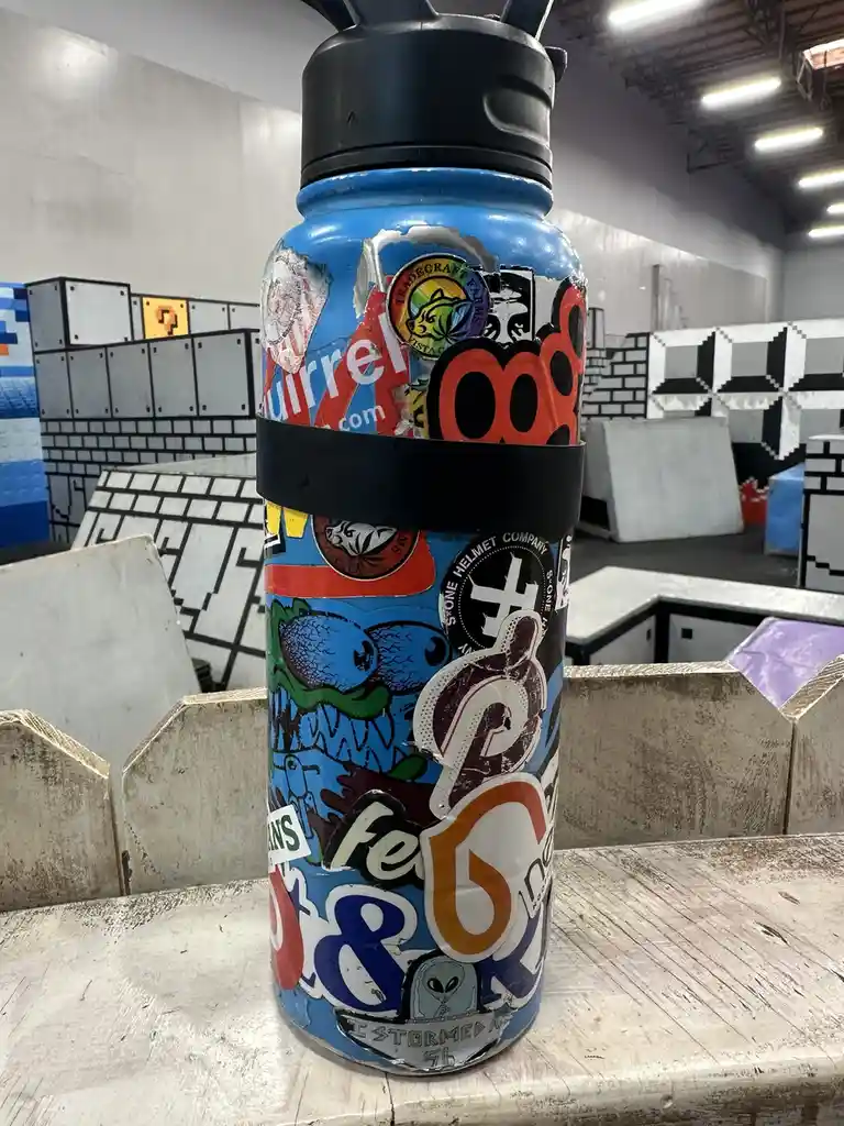 A blue water bottle covered in numerous peeling and overlapping stickers sits on a wooden surface in what appears to be an…