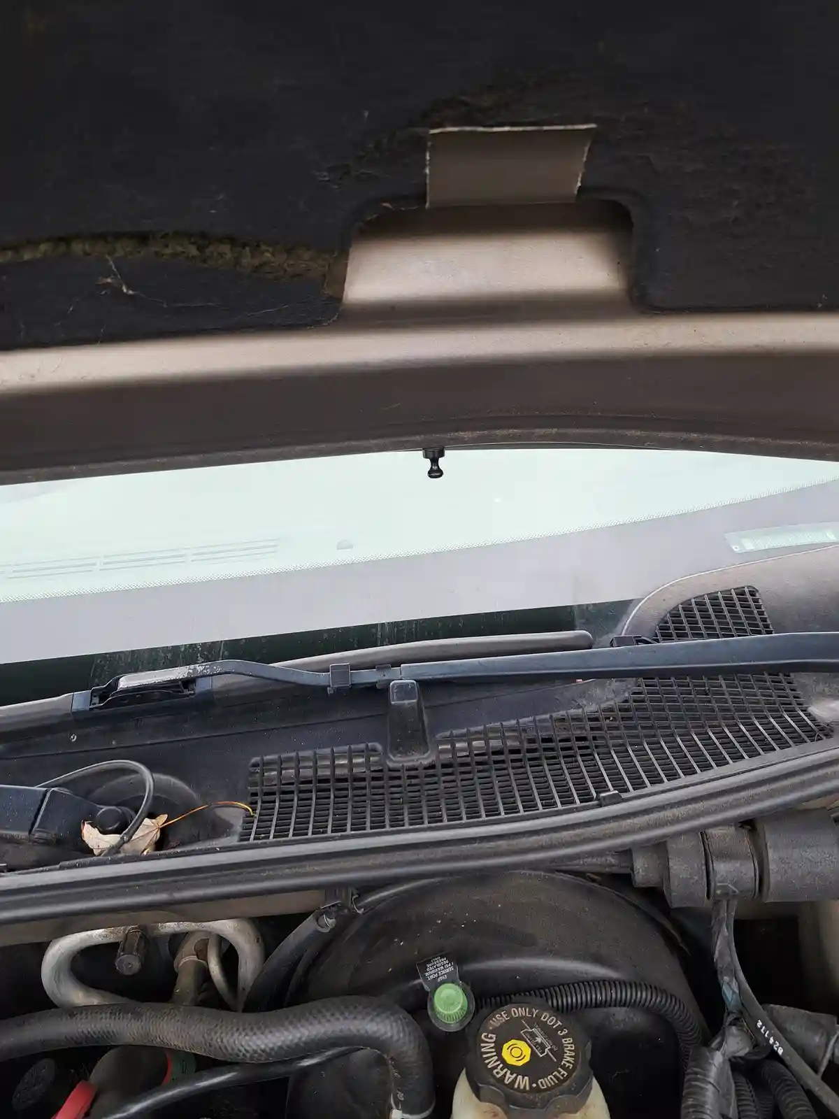 A close-up photo of an open car hood showing the engine bay, windshield, and wipers. The hood's inner lining is visibly torn…