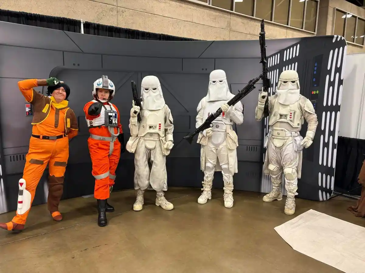 Five people in Star Wars costumes pose together: two in orange pilot gear, three in white First Order Stormtrooper armor…
