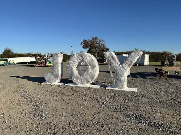 Three large, white, capital letters spelling 'JOY' are wrapped in clear plastic and sit on a gravel lot under a clear blue…