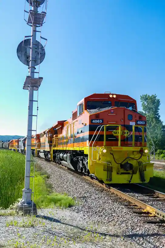 A bright orange and yellow NEORR locomotive numbered 4063 is shown on a sunny day, approaching the viewer on a gravel…