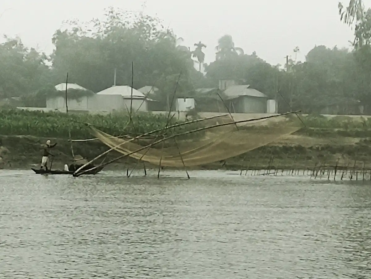 A fisherman in a small boat is casting a large, wide fishing net into a river. The net is suspended by bamboo poles and…