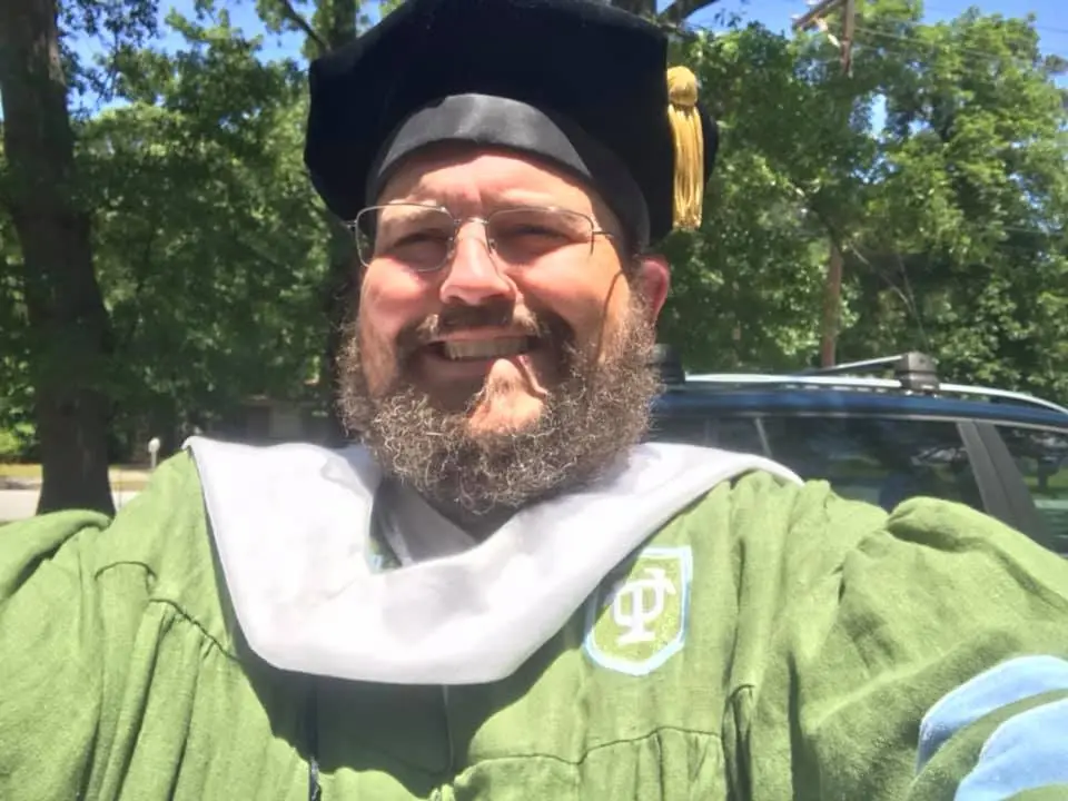 A man with a beard and glasses wears a green graduation gown and black cap with a gold tassel, smiling broadly at the camera…