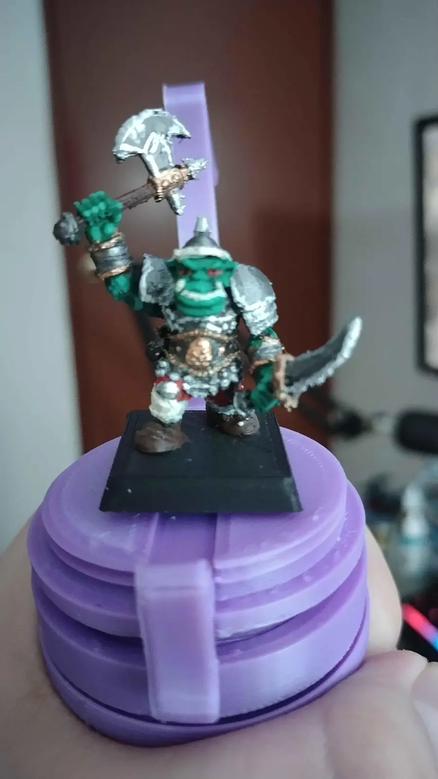 A green-skinned, armored fantasy orc miniature holding a large axe and a dagger stands on a black base, which is mounted…