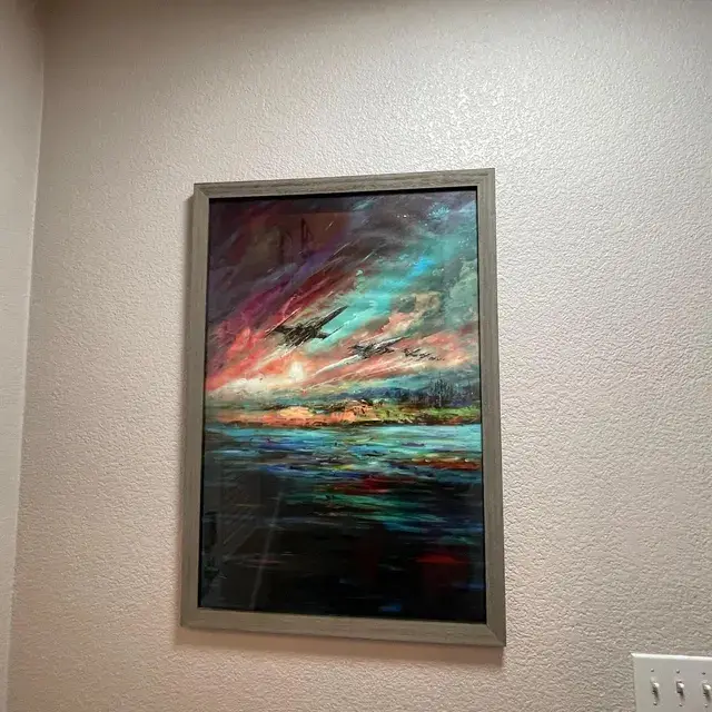 A framed painting hangs on a textured wall, depicting a dramatic sky with streaks of pink, orange, and teal, over a dark…