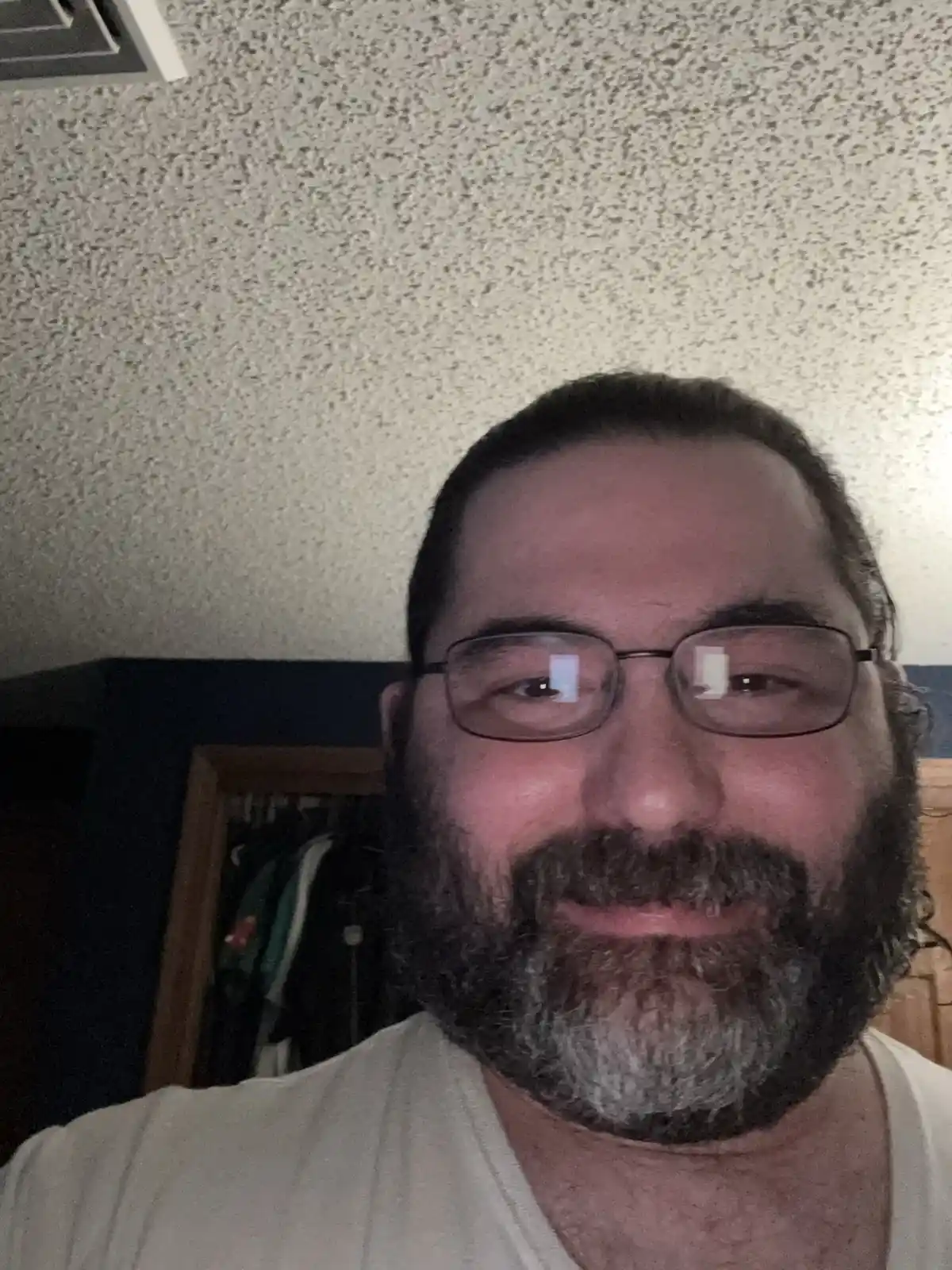 A close-up selfie of a middle-aged man with a full beard showing gray hairs, wearing glasses and a white t-shirt, smiling…