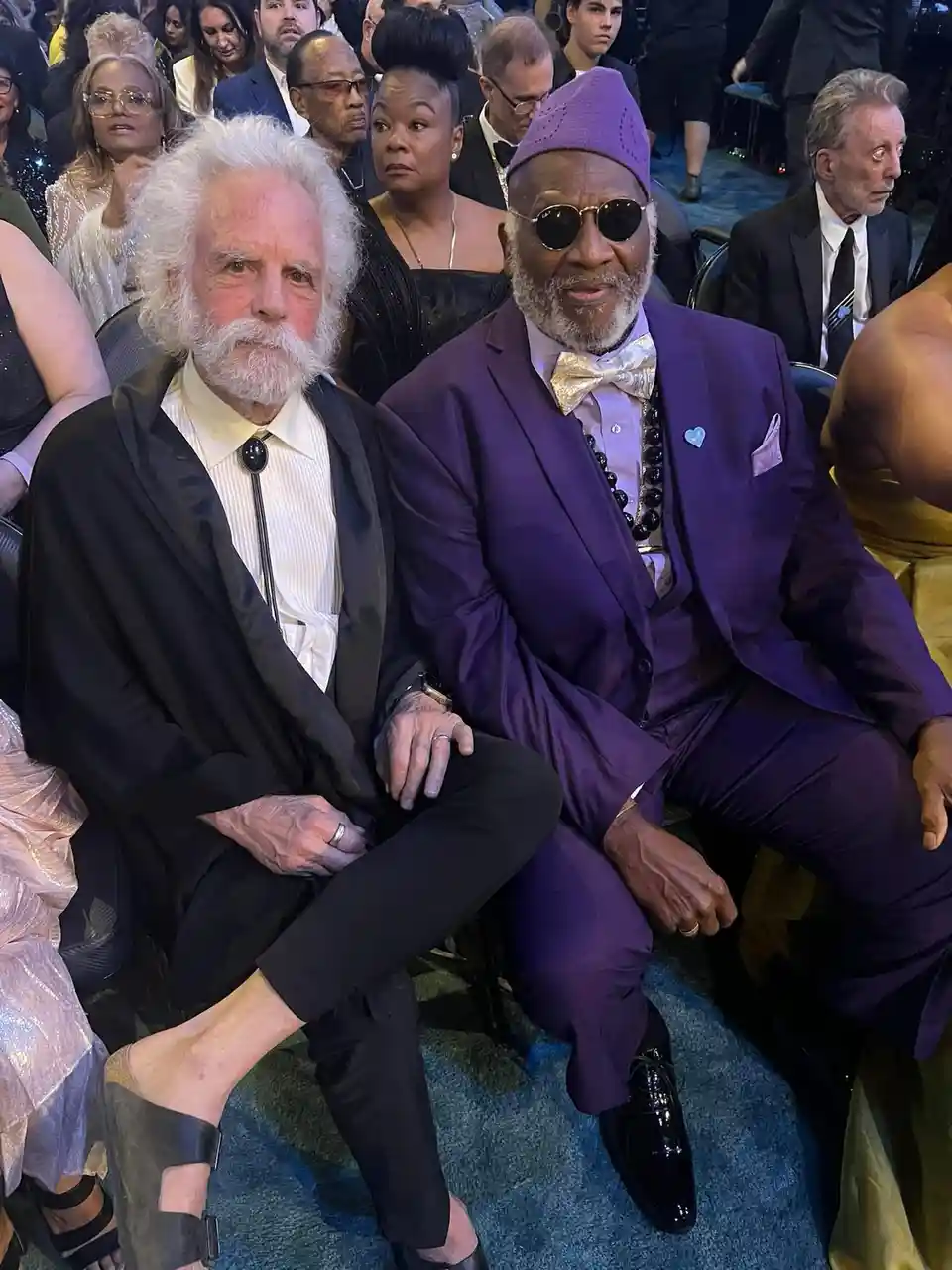 Two men sit side-by-side in a crowded, formal event. The man on the left has white hair and beard, wearing a black suit with…