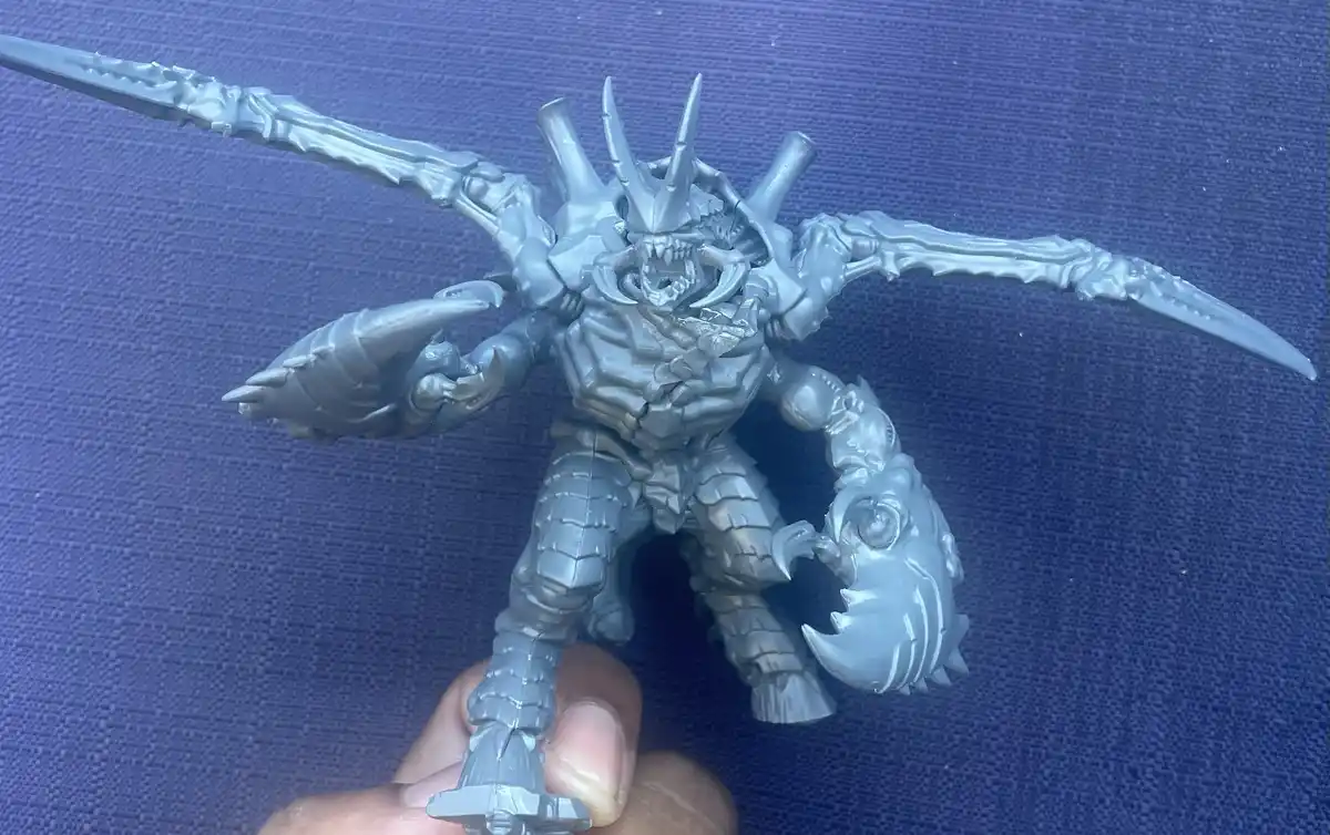 A person holds a gray, unpainted action figure of a monstrous, armored creature with multiple arms, claws, and spiked wings…