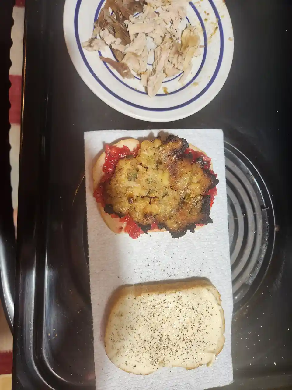 An overhead photo shows a partially assembled sandwich on a paper towel on a stovetop. The bottom bun is spread with butter…