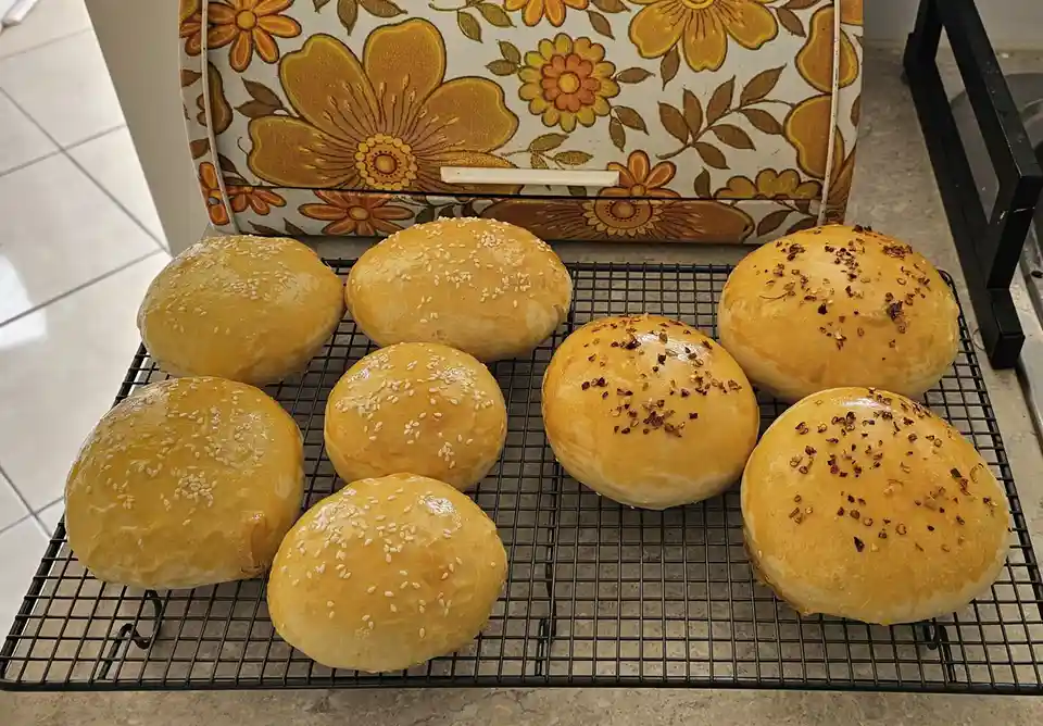 Eight freshly baked golden-brown burger buns sit on a black wire cooling rack. Four buns are topped with white sesame seeds…