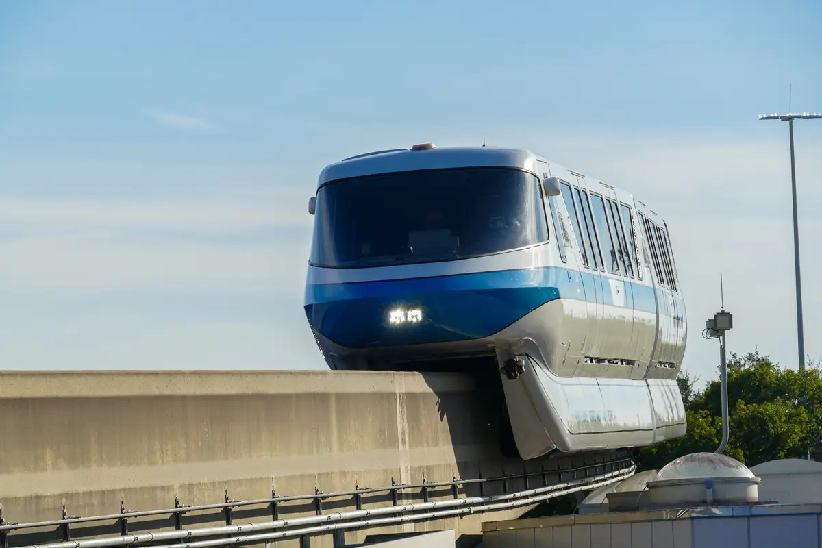 A blue and silver monorail train is shown approaching on an elevated track against a clear blue sky. The train is positioned…