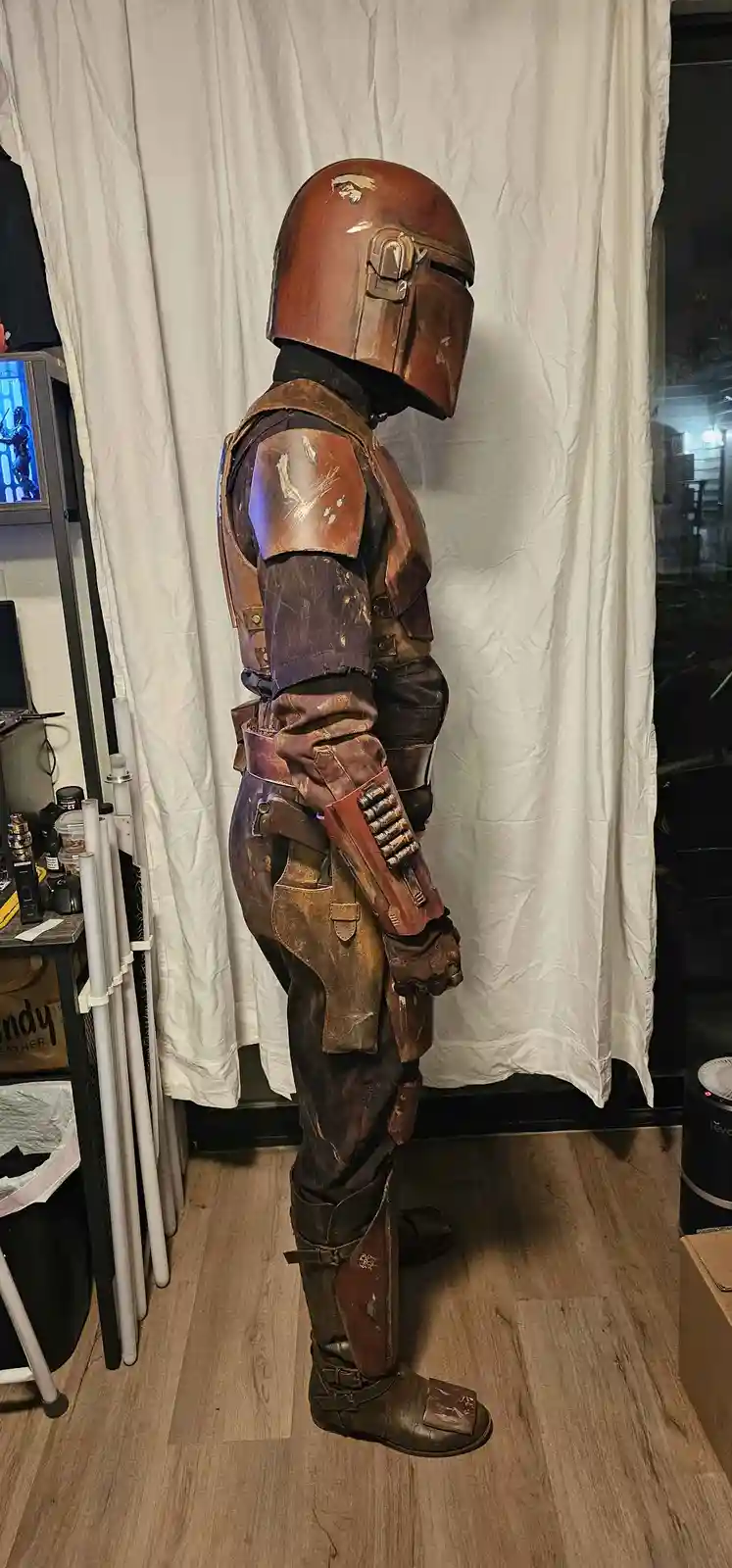 A person in a detailed, weathered Mandalorian armor costume stands in profile against a white curtain, holding a blaster…
