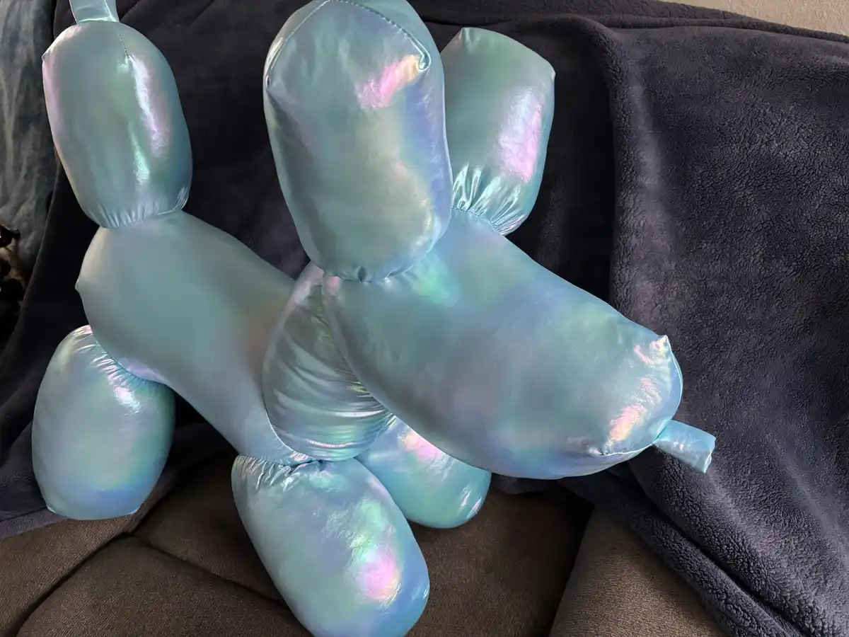 A plush toy shaped like a balloon dog, crafted from iridescent fabric that shifts between blue, green, and pink hues, rests…