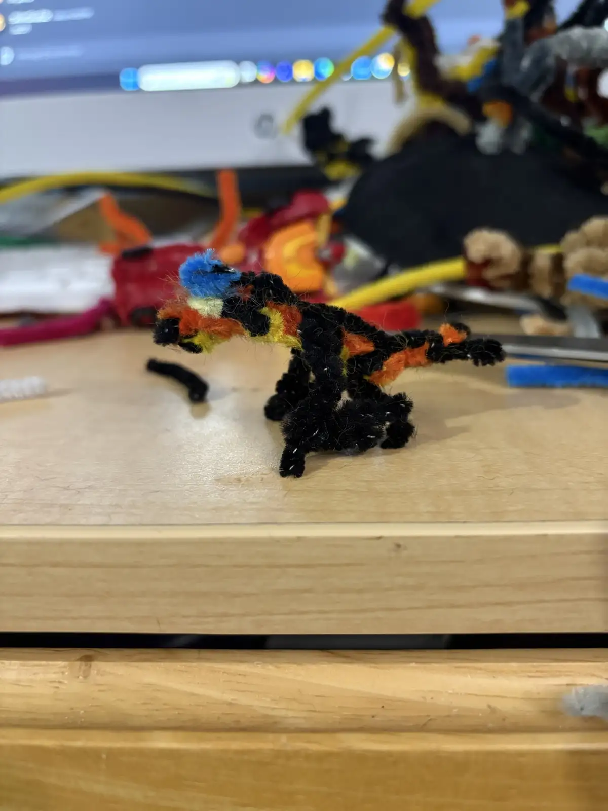 A small, handmade dragon figurine crafted from colorful pipe cleaners sits on a wooden desk, surrounded by crafting supplies…
