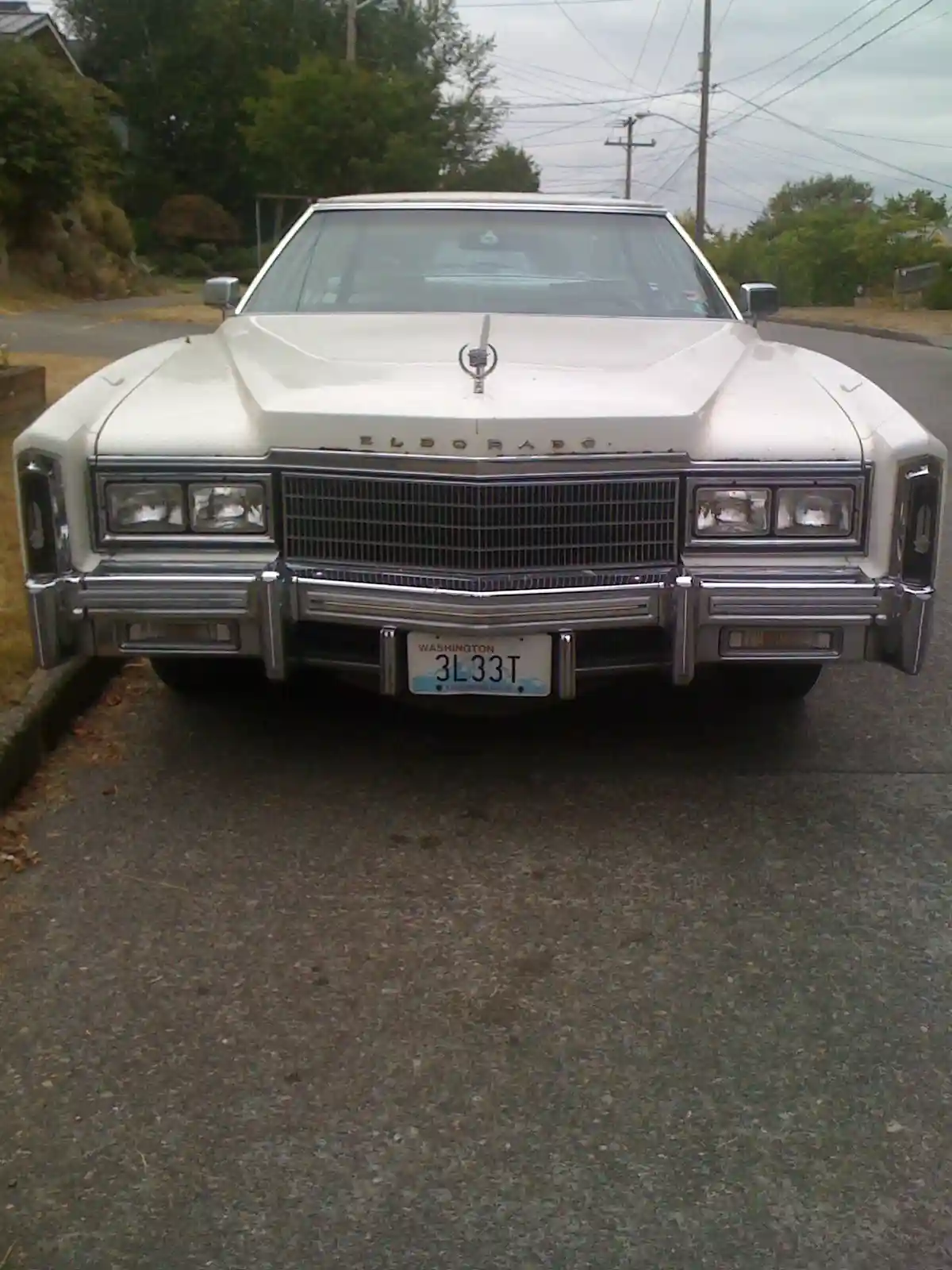A front-facing photo of a white 1970s Cadillac Eldorado with chrome grille and bumper, parked on a residential street. The…