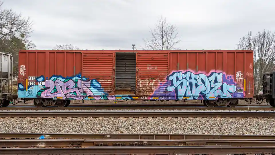 A red freight train car sits on tracks with its side doors open, covered in colorful graffiti. The overlaid text reads…