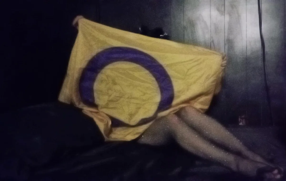 A person wearing fishnet stockings is sitting on a dark surface, covered by a yellow cloth with a blue circle, obscuring…