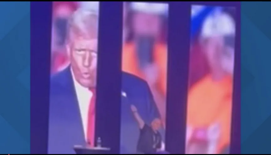 A blurry image shows a man in a suit speaking at a podium, with large vertical screens behind him displaying a close-up of…