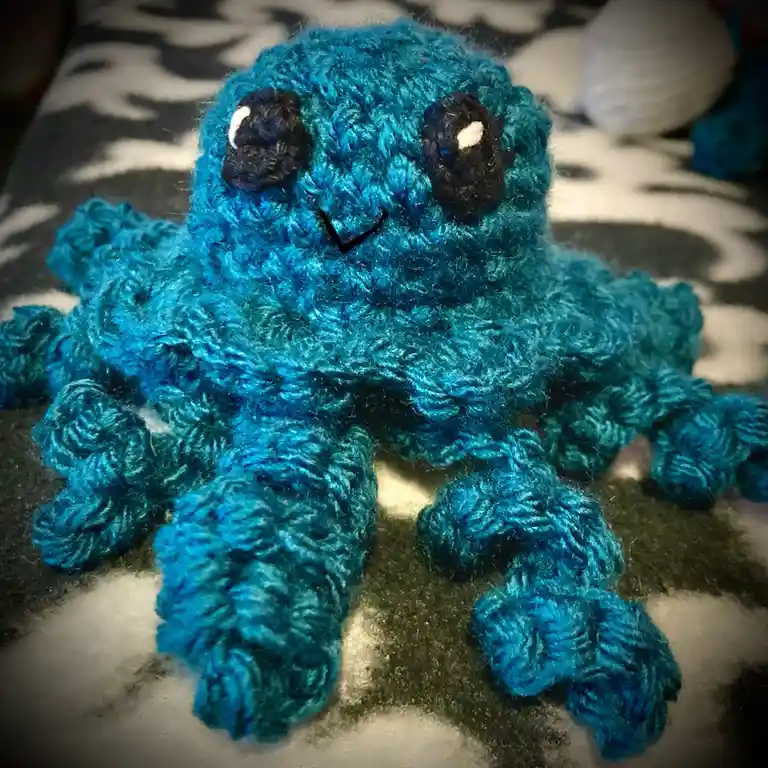 A handmade, blue crocheted octopus plush toy with large black eyes featuring white pupils and a simple black V-shaped smile…