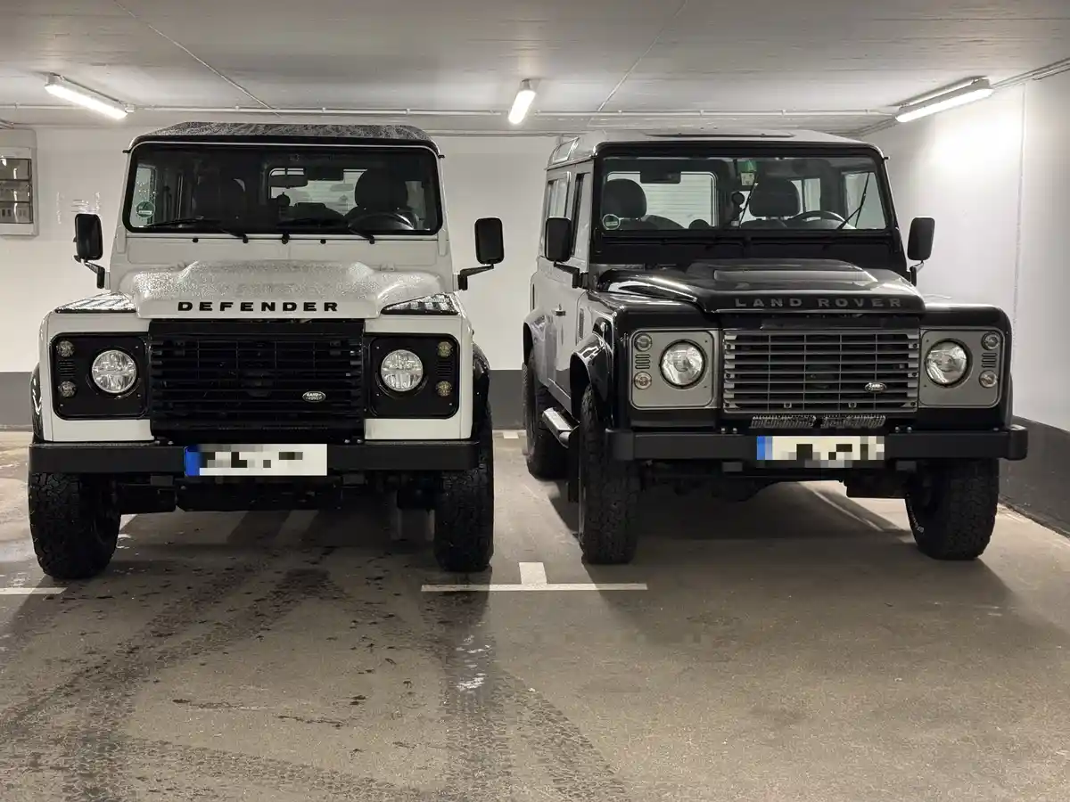 Two Land Rover Defender SUVs, one white and one black, are parked side-by-side in an underground parking garage. The white…
