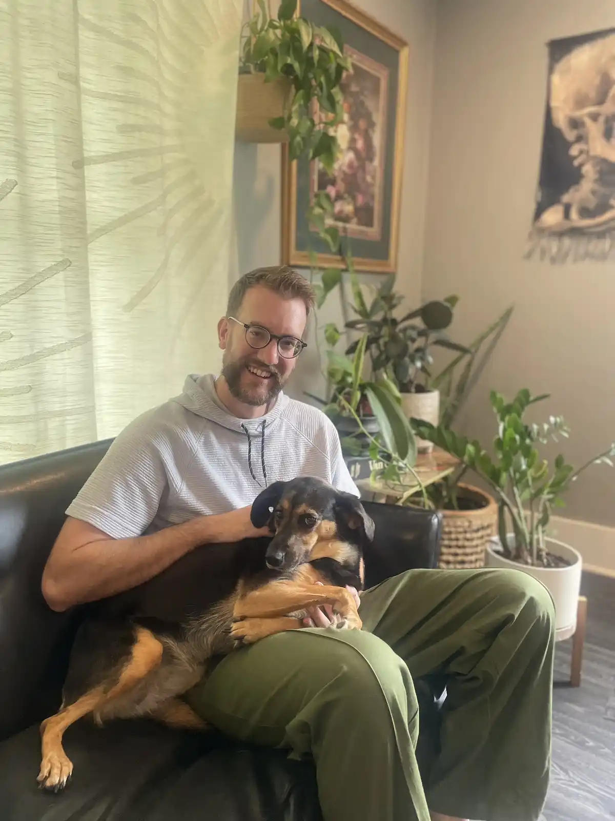A smiling man with glasses sits on a black couch holding a black and tan dog that looks unimpressed. The room is decorated…