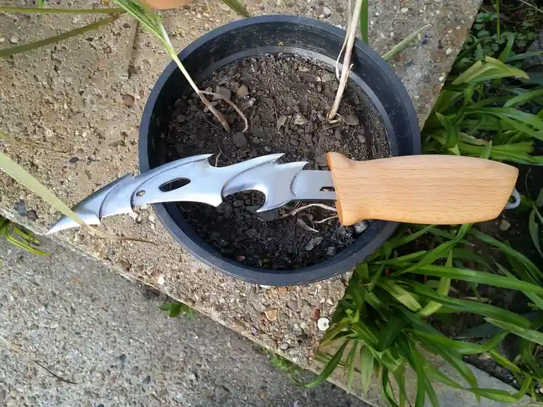 A decorative, ornate garden trowel with a wooden handle and a silver, bat-like blade design is stuck in a black plastic pot…