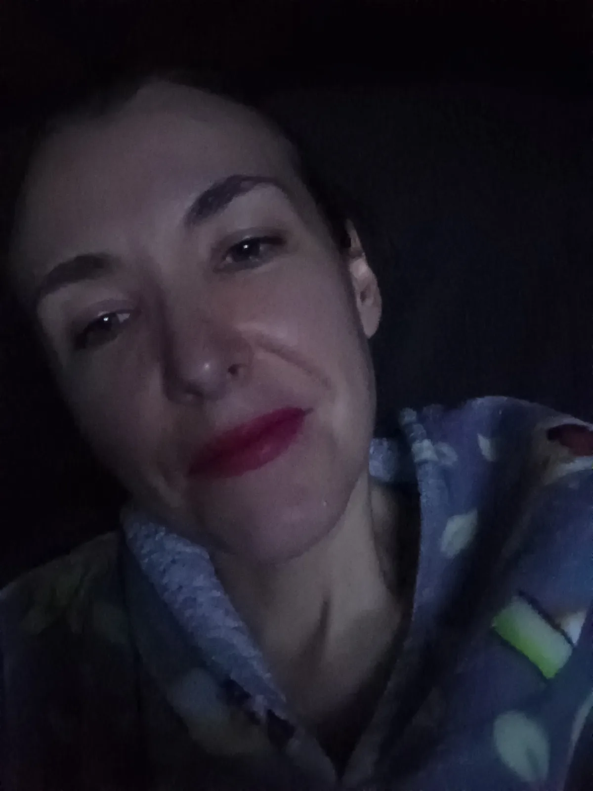 A person with red lipstick lies in a dark room wearing a patterned pajama top, looking directly at the camera with a tired…