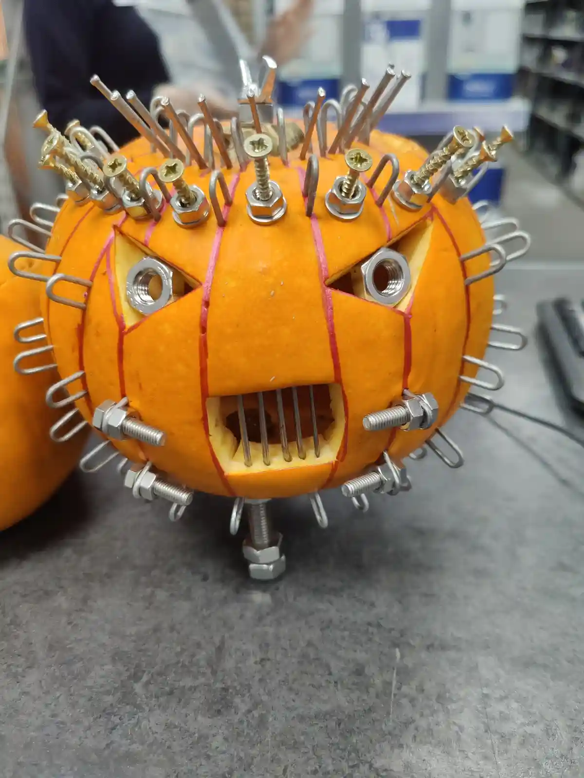 a close-up of an orange pumpkin carved into a face with angry eyes and a mouth, decorated with numerous screws, bolts, and…