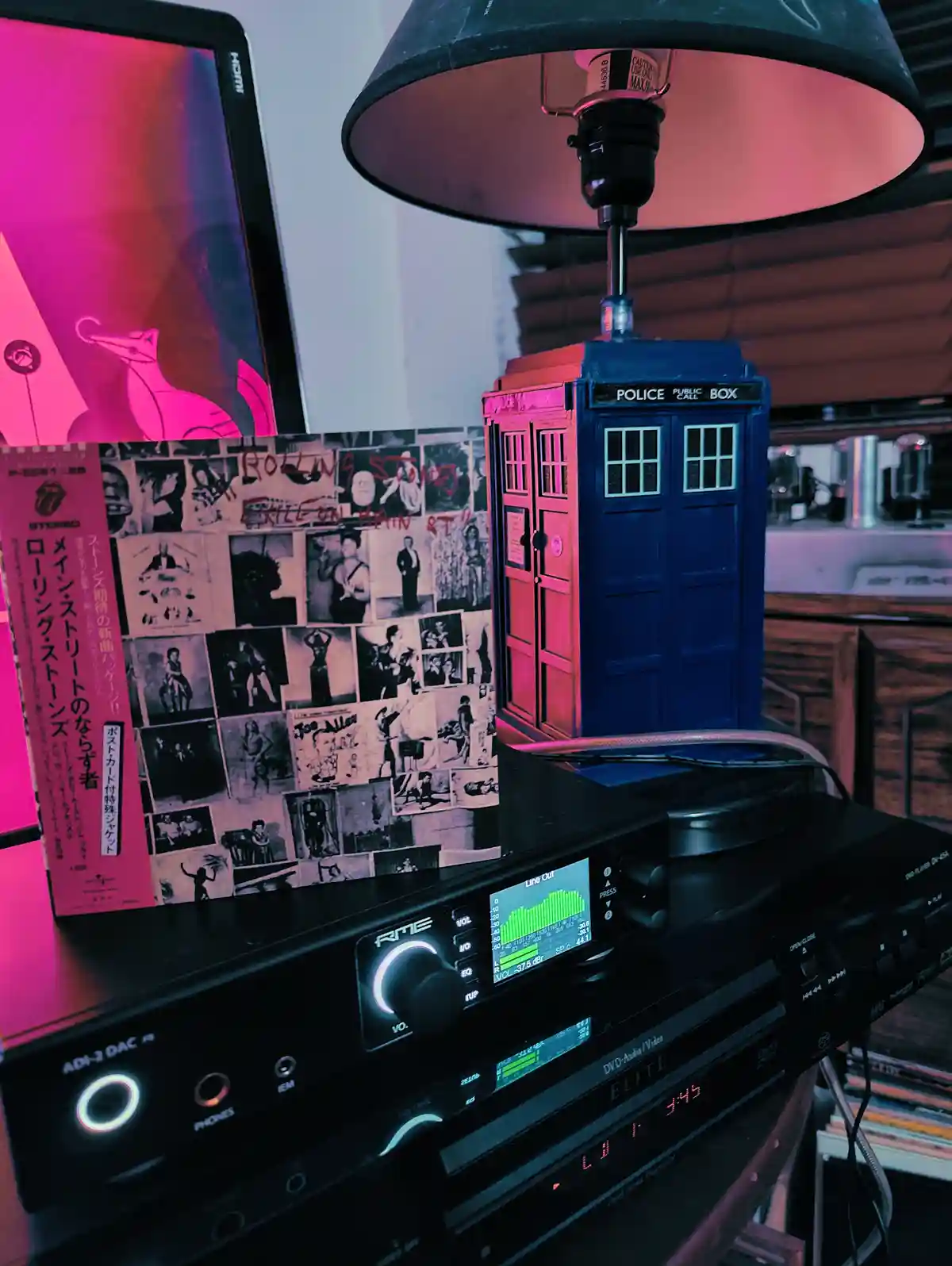 A blue police box lamp sits atop a black audio system, surrounded by vinyl records and a laptop displaying pink light. The…