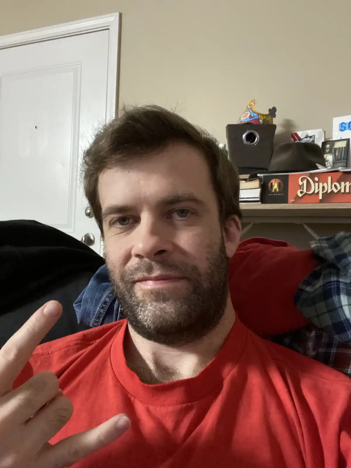 A man with a beard and red shirt makes a peace sign while looking at the camera, with a shelf behind him holding board games…