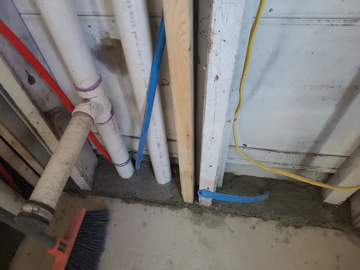 A close-up photo of a messy basement or utility area with white PVC pipes, a blue electrical cable, a yellow cable, wooden…
