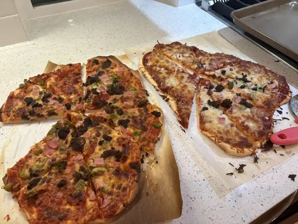 Two rectangular pizzas, sliced into triangles, sit on parchment paper on a kitchen counter. Both pizzas have cheese, ham…