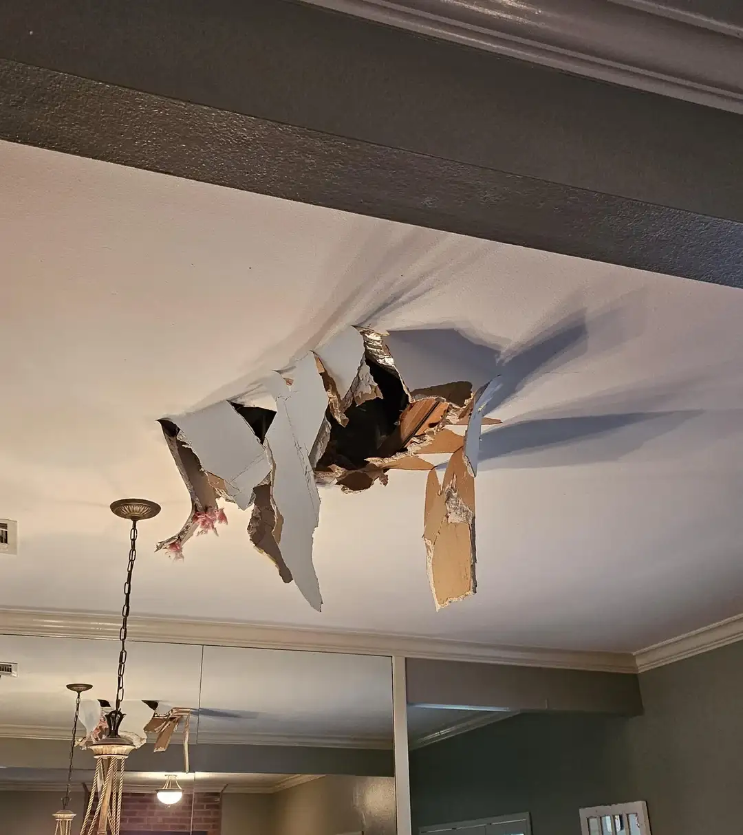 A large, jagged hole rips through a white ceiling, exposing the brown insulation and structural layers beneath. The ceiling…