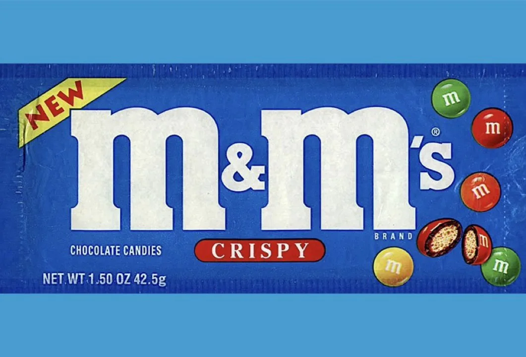 A blue wrapper for a new M&M's Crispy chocolate candy product, featuring the brand logo, a 'NEW' banner, and images of…