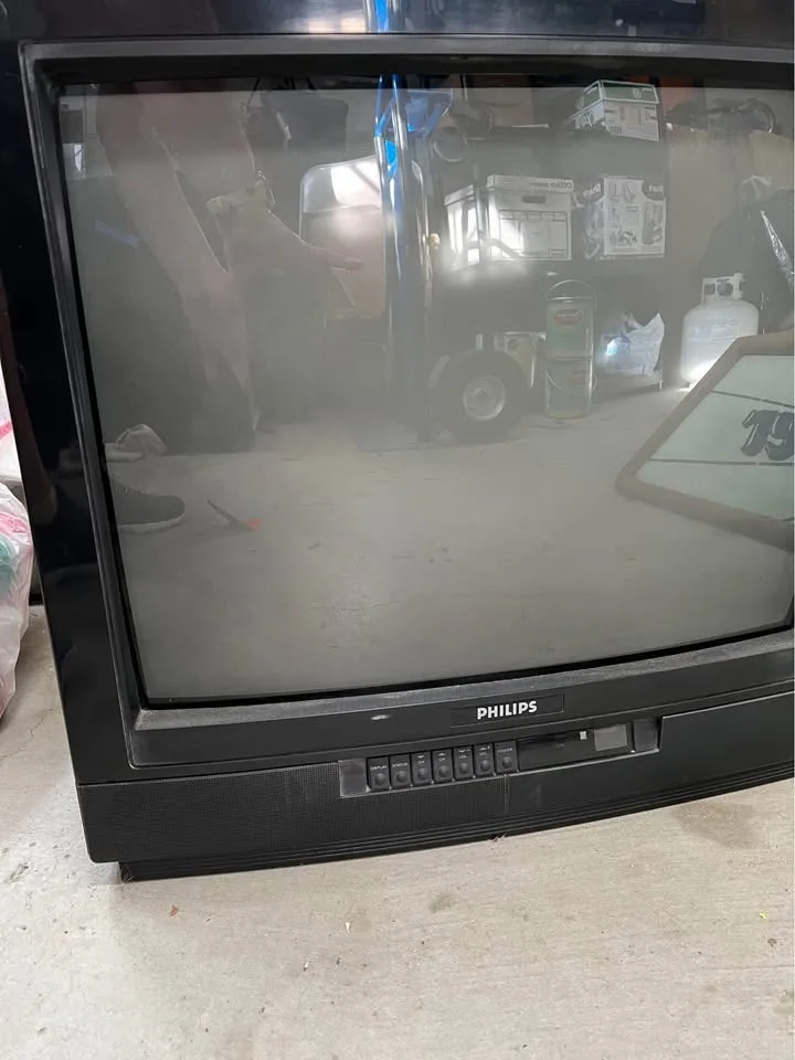 A dusty, old black Philips CRT television sits in a cluttered garage, its screen blank and smudged with grime. The…