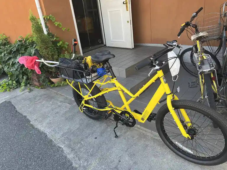 a bright yellow bicycle is parked outside a building, heavily modified with a large rear rack holding a black bag, a yellow…