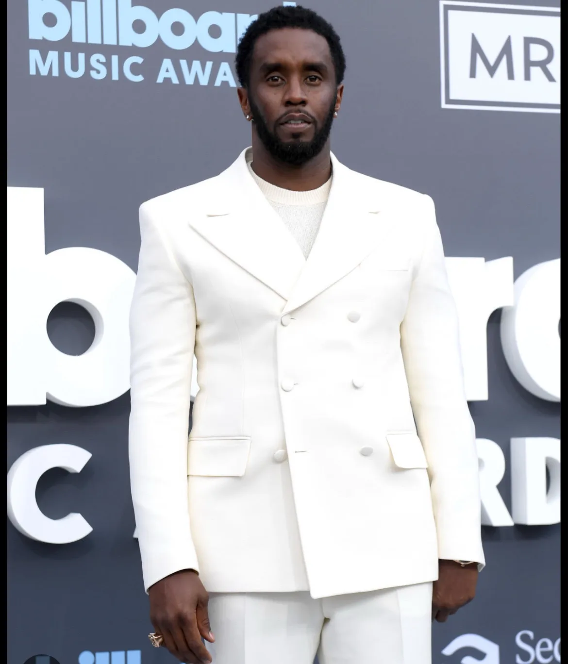 A man with a beard and short hair, wearing a white double-breasted suit and a light-colored sweater, stands on a red carpet…
