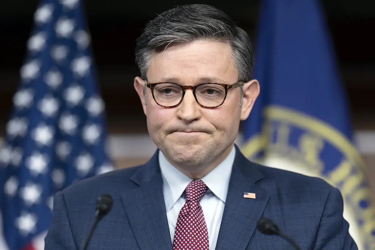 A man in a dark suit, white shirt, and red patterned tie stands at a podium with microphones, wearing tortoiseshell glasses…