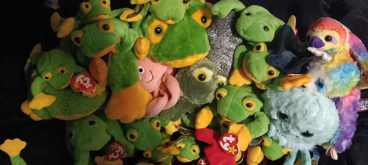A large pile of plush toys, mostly green frogs with yellow feet and eyes, mixed with a few other colorful stuffed animals…