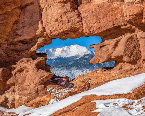 A scenic photograph shows a natural red rock arch framing a distant snow-capped mountain under a clear blue sky. Snow…