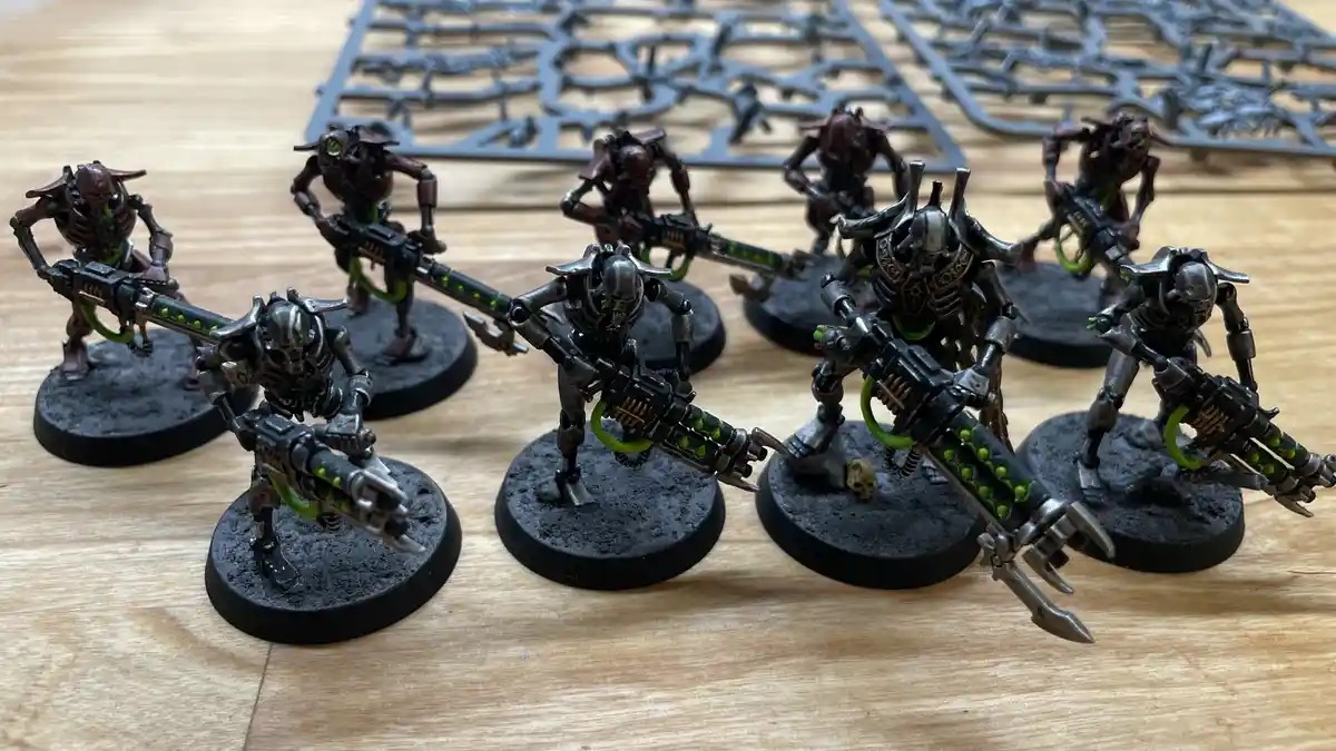 A row of painted miniature sci-fi soldiers with skull-like helmets and large green-accented weapons stand on black circular…