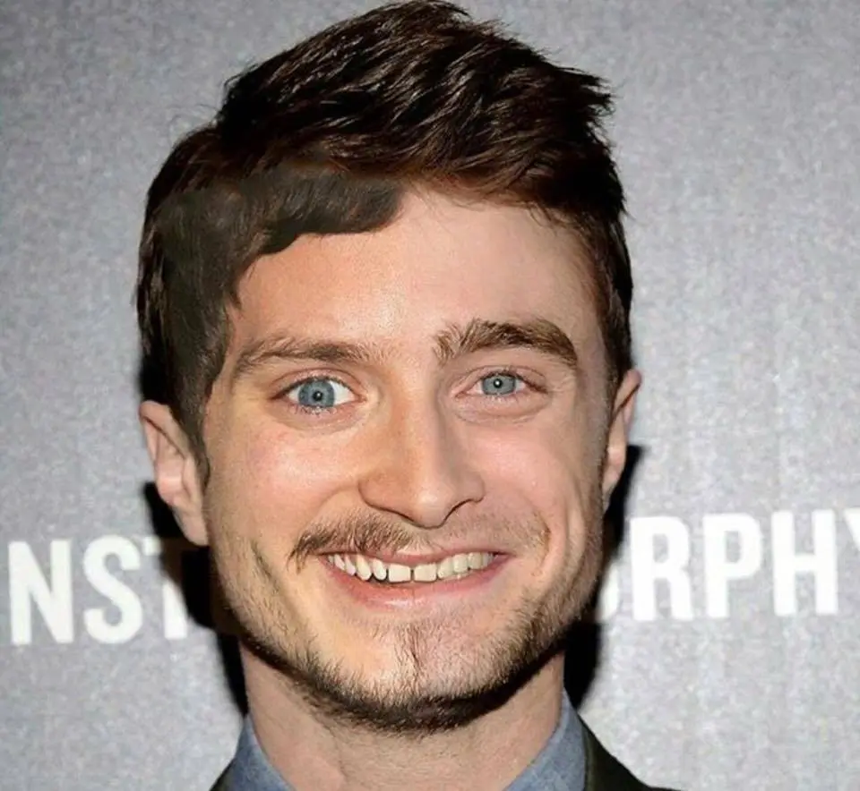 A close-up photo of actor Daniel Radcliffe smiling broadly with his teeth showing, against a gray backdrop with partially…