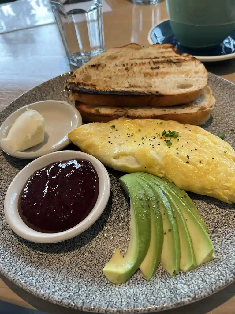 A plate of breakfast food including a folded omelette, sliced avocado, grilled toast, a small dish of butter, and a small…