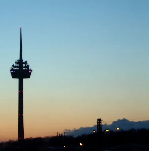 A silhouette of a tall, slender tower with a pointed top and a lit observation deck against a twilight sky, with faint city…