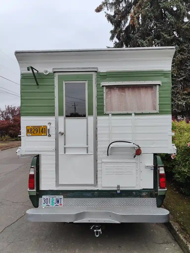 The image shows the rear of a vintage green and white camper trailer parked on a street, featuring a door, a window with a…