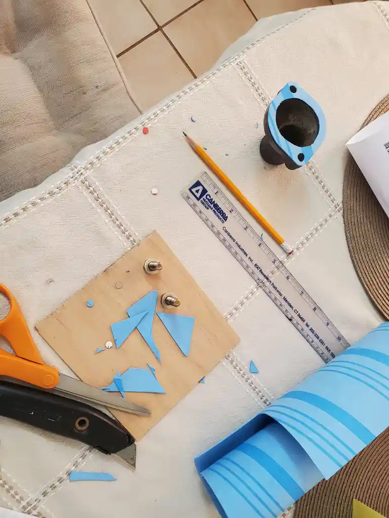 An overhead shot shows a beige cushion with various crafting tools: orange-handled scissors, a black utility knife, a yellow…