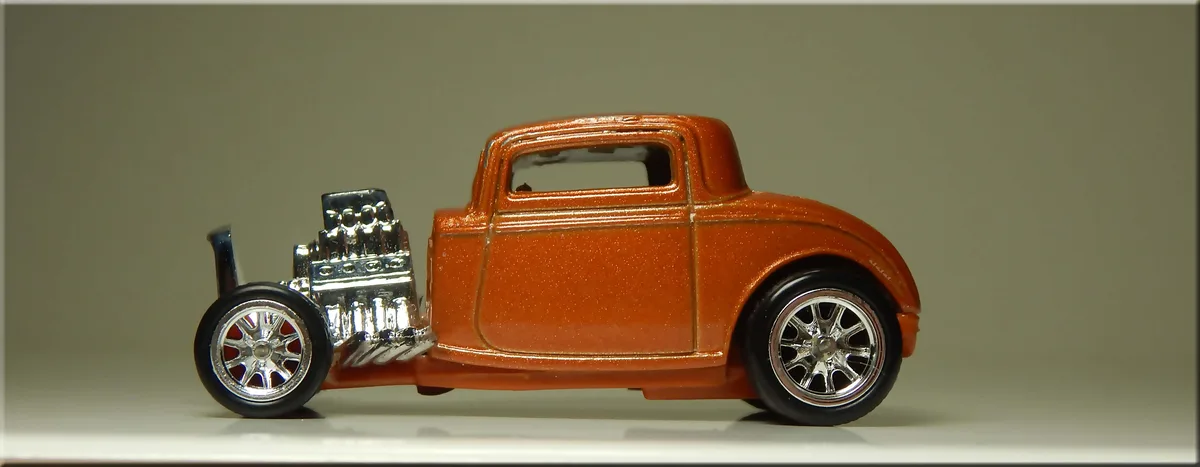 A side-profile photo of a detailed orange Hot Wheels-style toy hot rod with a chrome engine and wheels, set against a plain…