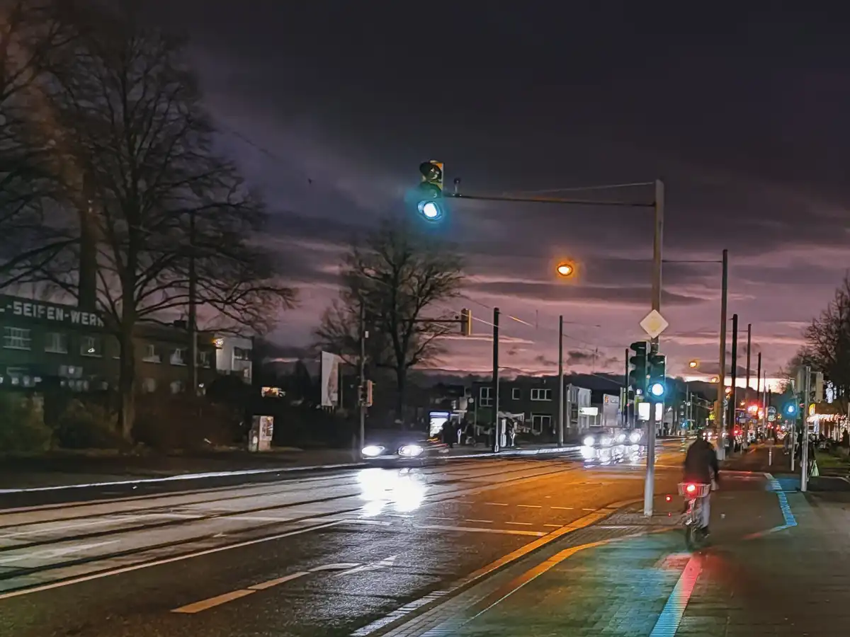 A nighttime street scene with wet pavement reflecting streetlights and car headlights, a cyclist rides on a bike path next…